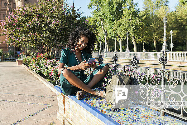 Smiling female tourist using mobile phone while sitting cross-legged on bench at Plaza De Espana  Seville  Spain