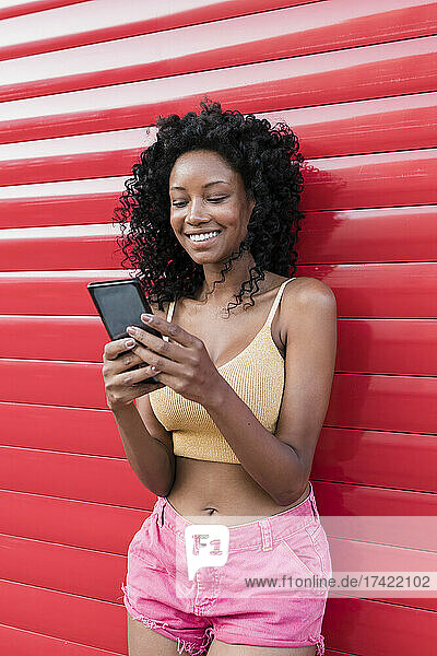 Smiling woman with curly hair using smart phone in front of red shutter
