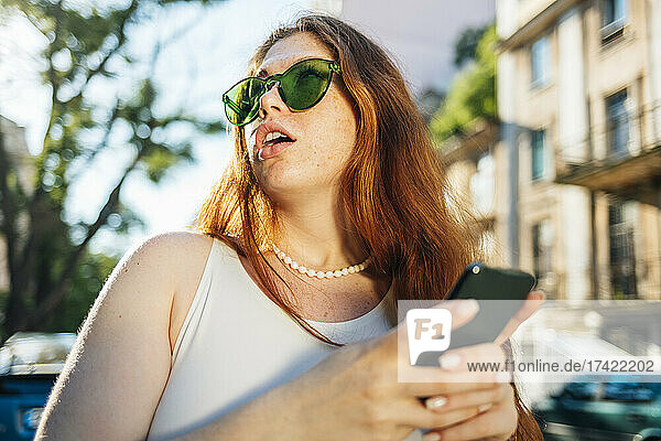 Surprised young woman in green sunglasses holding mobile phone