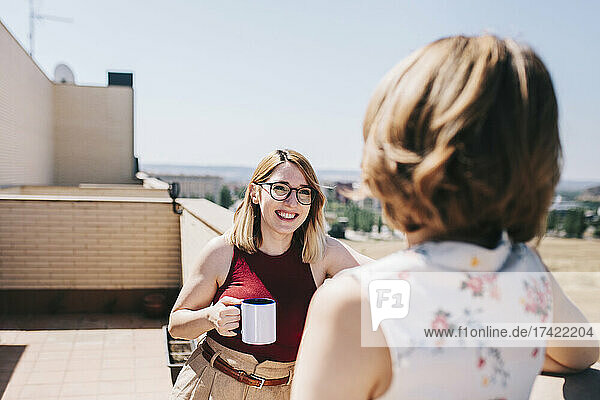 Fröhliche Geschäftsfrau hält Kaffeetasse in der Hand  während sie mit einem Kollegen auf dem Dach diskutiert