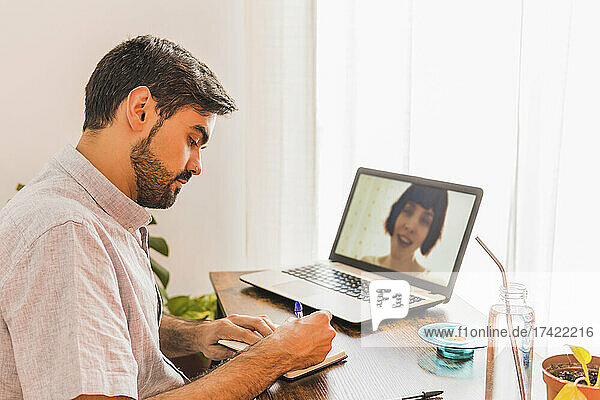 Male doctor taking notes while listening to female patient on video call through laptop