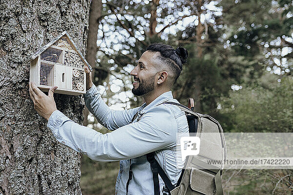 Smiling male backpacker hanging insect hotel on tree in forest