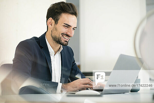 Happy young businessman using laptop while sitting at desk in office