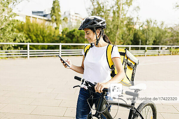 Smiling delivery woman using mobile phone while wheeling bicycle