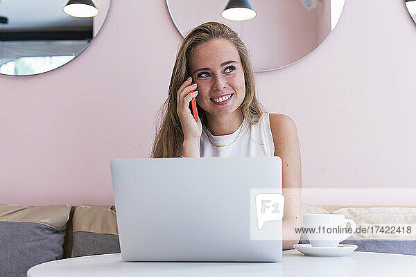 Smiling female freelancer talking on mobile phone while sitting with laptop in cafe