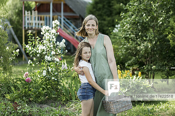 Mother embracing daughter while holding wicker basket in backyard