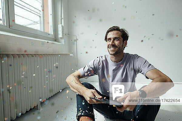 Man with digital tablet looking at confetti while sitting on floor
