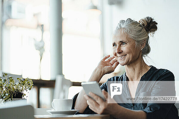Smiling mature female freelancer with mobile phone sitting in coffee shop