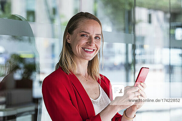 Smiling businesswoman holding mobile phone in front of glass wall