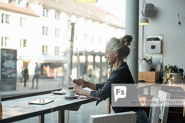 Female freelancer using mobile phone while sitting in cafe by glass window