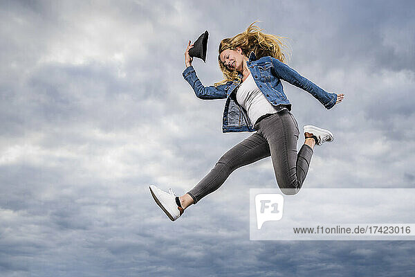 Young woman jumping against cloudy sky with hat in hand