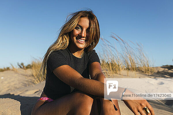 Glückliche Frau mit braunen Haaren  die an einem sonnigen Tag auf Sand sitzt