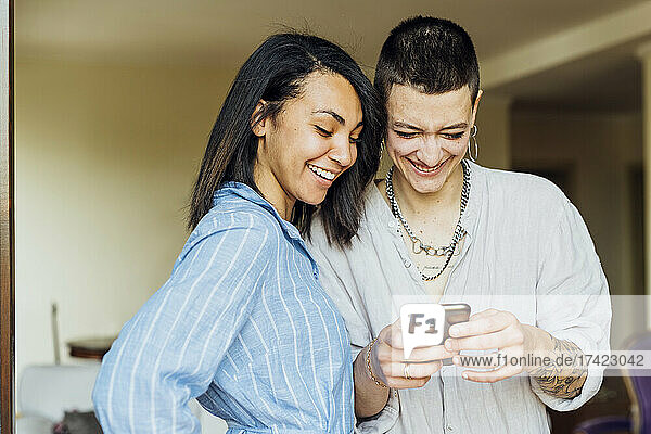 Smiling young woman using phone by girlfriend at home