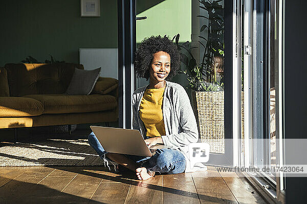 Lächelnde junge Frau mit Laptop sitzt zu Hause im Sonnenlicht