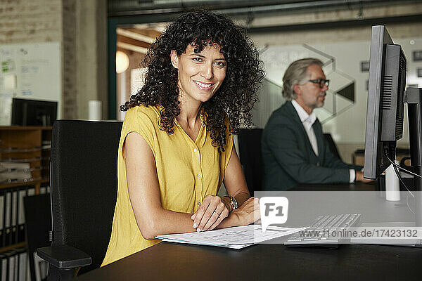 Smiling female professional with documents sitting at desk in office