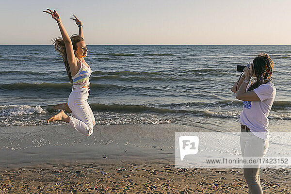 Woman photographing female friend jumping at beach