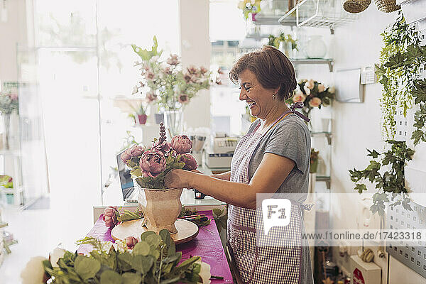 Fröhliche reife Floristin arrangiert Blumen  während sie am Tisch im Geschäft steht