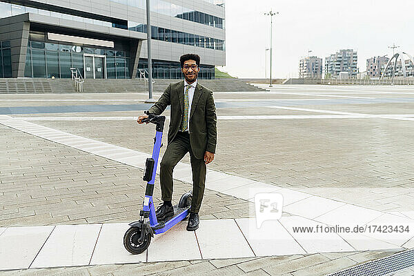 Smiling businessman standing with electric push scooter on footpath