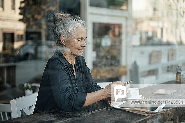 Female freelancer using mobile phone while sitting in cafe seen through glass window