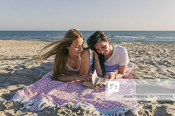 Young women reading book while lying on picnic blanket at beach