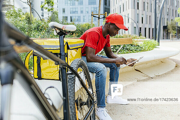 Young delivery man using digital tablet while sitting by package on bench
