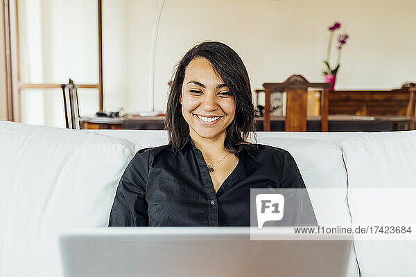 Lächelnde junge Frau benutzt Laptop  während sie zu Hause auf dem Sofa sitzt