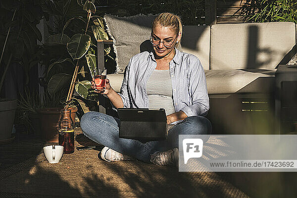 Mature businesswoman holding drink while sitting with digital tablet on terrace