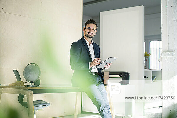 Handsome young businessman holding digital tablet while sitting on desk in office