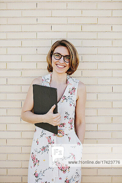 Lächelnde Geschäftsfrau mit Brille und digitalem Tablet steht vor einer Ziegelwand