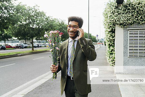 Smiling businessman holding bunch of flowers while talking on mobile phone on street