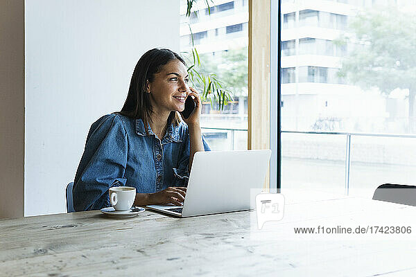 Smiling businesswoman talking on mobile phone while sitting with laptop in cafe