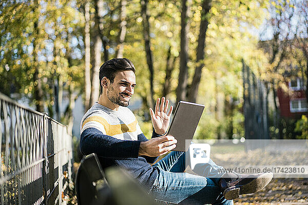 Young man waving hand while doing video call through digital tablet at park during sunny day