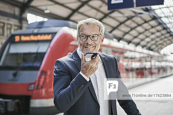 Lächelnder männlicher Berufstätiger  der wegschaut  während er am Bahnhof Voicemail über sein Smartphone sendet