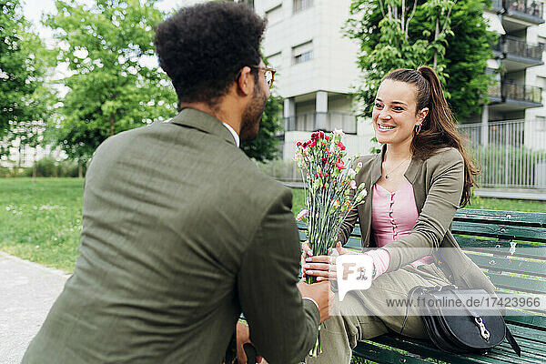 Smiling businesswoman taking bouquet from colleague while sitting on park bench
