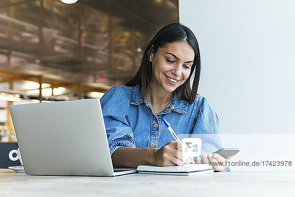 Smiling businesswoman writing on diary while holding mobile phone at coffee shop