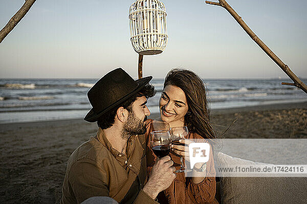Lächelndes Paar hält Getränke in der Hand  während es am Strand sitzt