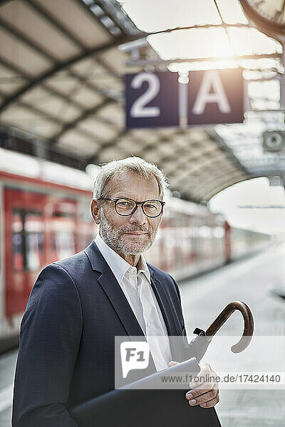 Senior businessman with laptop and umbrella standing on railroad platform
