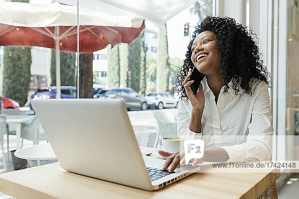Happy businesswoman with laptop talking on smart phone at cafe