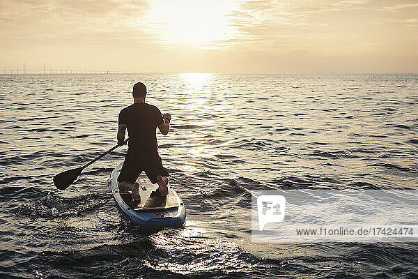Rear View of man rowing paddleboard in sea during sunset