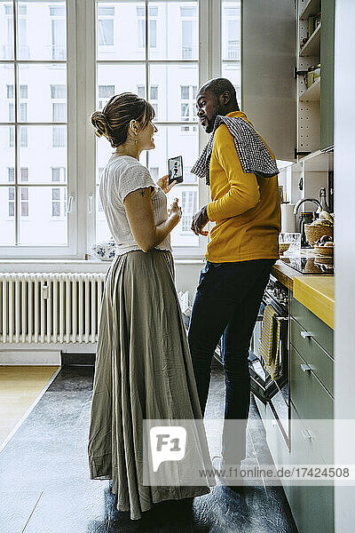 Lächelnde Frau mit Smartphone im Gespräch mit ihrem Freund bei der Hausarbeit in der Küche in voller Länge