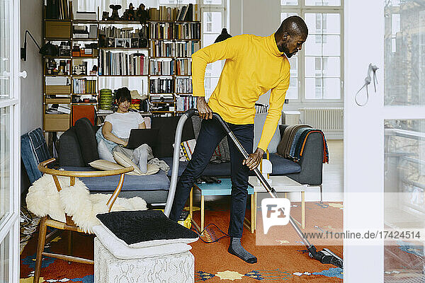 Mann reinigt Teppich in voller Länge mit Staubsauger  während eine Freiberuflerin im Wohnzimmer zu Hause am Laptop arbeitet