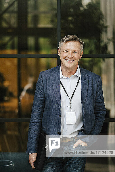Portrait of smiling mature businessman standing with hand in pocket in front of glass wall at convention center