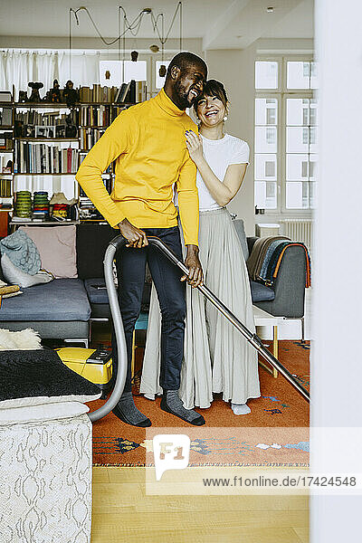 Fröhlicher Mann benutzt Staubsauger in voller Länge  während er neben einer Frau im Wohnzimmer steht