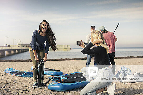 Glückliche Frau  die ein Paddleboard aufpumpt  während eine Freundin am Strand fotografiert