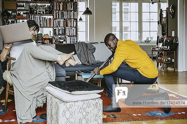 Mann reinigt Teppich in voller Länge mit Staubsauger  während Frau mit Laptop auf einem Stuhl im Wohnzimmer zu Hause sitzt