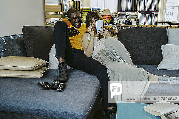 Paar in voller Länge  das lacht  während es ein Smartphone benutzt und zusammen auf dem Sofa im Wohnzimmer sitzt
