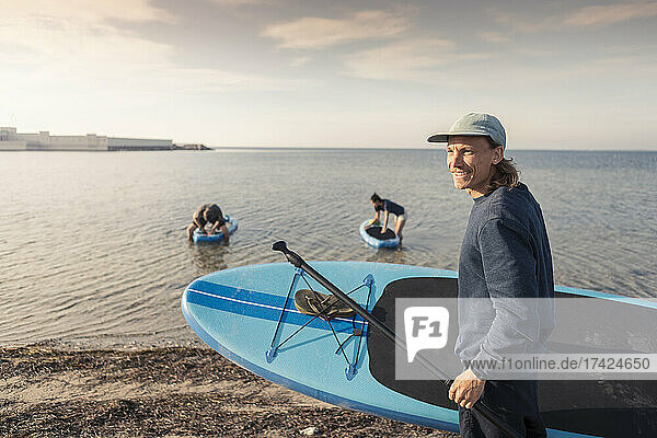 Männlicher Lehrer mit Paddleboard am Strand bei Sonnenuntergang