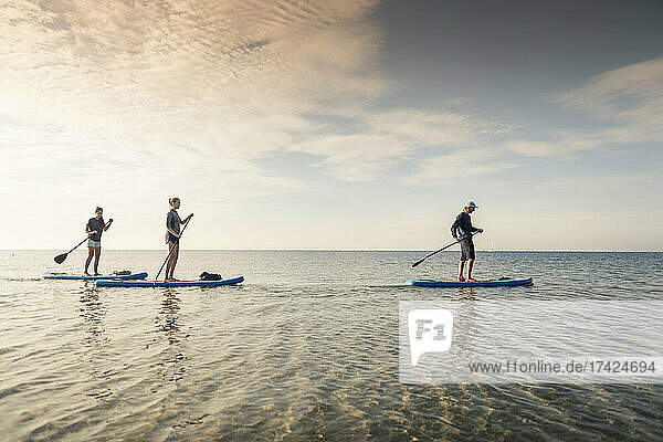 Weibliche Freunde lernen Paddleboarding von einem männlichen Lehrer im Meer bei Sonnenuntergang