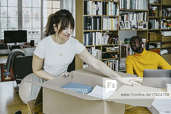 Mid adult woman opening cardboard box by man sitting with laptop at table
