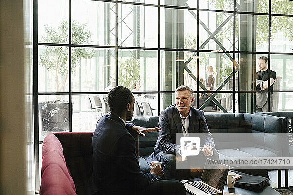 Reifer Geschäftsmann schaut auf einen männlichen Fachmann  der auf einem Sofa im Kongresszentrum sitzt und diskutiert
